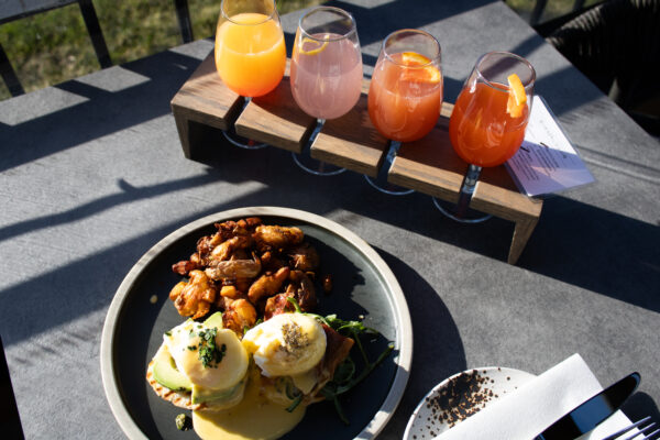Brunch and Mimosas at Cannon (14) A plated brunch dish of eggs Benedict with potatoes sits on an outdoor table alongside four colorful mimosas in a wooden tasting flight.