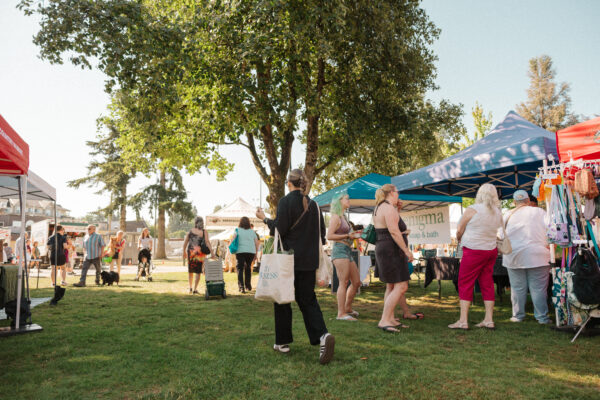 Abbotsford Farm & Country Market People browse vendor tents and displays at an outdoor market set on grass beneath leafy trees on a sunny day.