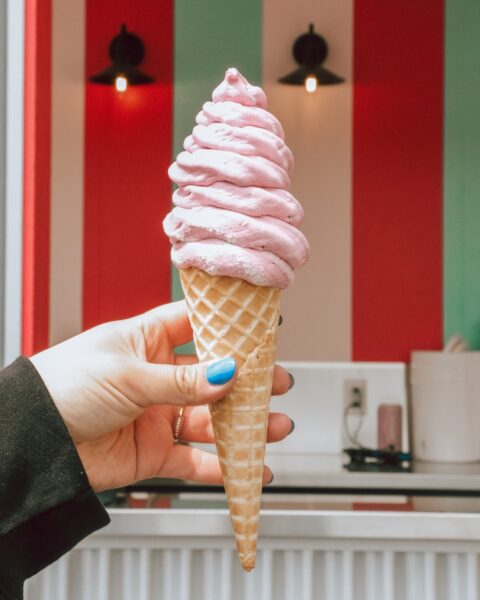 Lepp Ice Cream A hand holds a waffle cone topped with a tall swirl of pink ice cream inside a brightly striped ice cream shop.