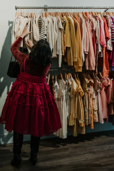 Photo Credit: Rachel Barkman A person reaches for clothing on a rack filled with neatly hung garments in neutral and pastel tones inside a boutique.