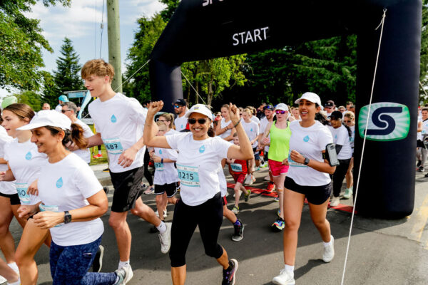 Run for Water – Alex Harte A large group of participants jog forward together as they cross a start line at an outdoor fun run event.